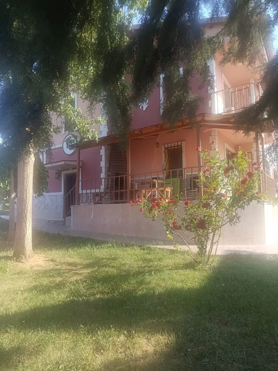 Ağaç gölgesinde çimenlik, kırmızı güller ve arkada villanın terası