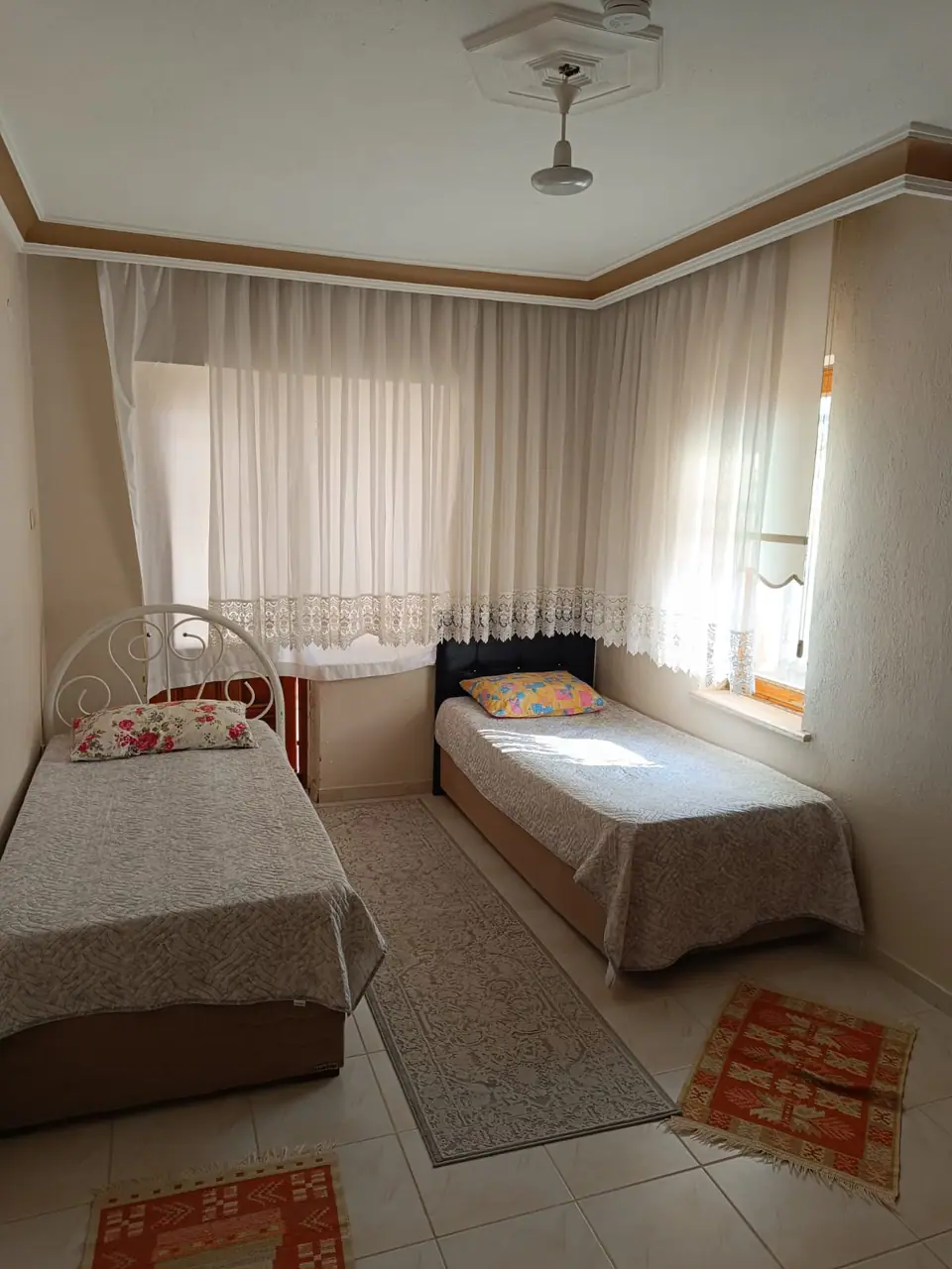Köşe pencereli, dantel perdeli ve iki tek kişilik yataklı aydınlık oda